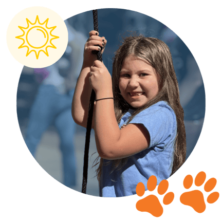 A smiling elementary-aged girl wearing a blue shirt holds onto a playground rope, looking toward the camera during outdoor play on a sunny day.