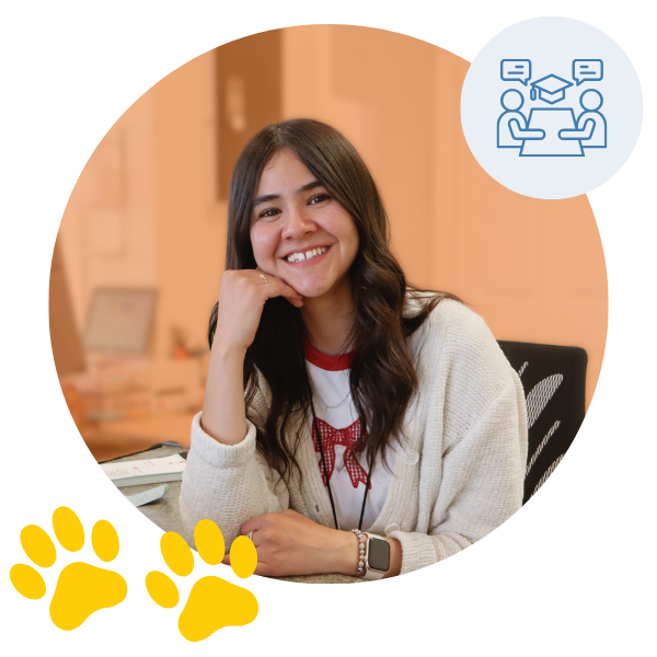 Smiling woman sitting at a desk in a classroom setting with a blue icon of two people at a table and two yellow paw prints, inside an orange-toned circular background. Smiling woman sitting at a desk in a classroom setting with a blue icon of two people at a table and two yellow paw prints, inside an orange-toned circular background.