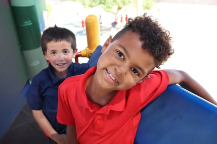 Two young students smile while standing together on playground equipment, with one student leaning toward the camera.